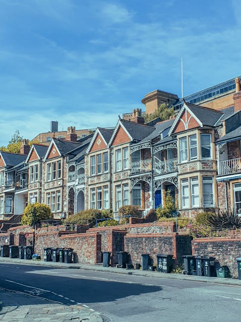 A row of Victorian-style terraced houses with decorative wrought iron balconies and detailed woodwork on the gabled roofs, set on a slope above a paved street. The houses are constructed with brick facades, some featuring white painted elements to highlight architectural details. In front of the properties, there is a low brick wall with small landscaped garden areas containing shrubs and small trees. Along the street, several black and green waste bins are aligned near the curb, indicating waste collection or disposal activity. The scene is illuminated by natural daylight, with a clear blue sky overhead, and a modern building with glass windows and a rooftop extension is visible behind the houses, providing a contrasting backdrop to the historic architecture. This scene exemplifies a typical residential area where private waste management services, such as rubbish removal, are often employed alongside traditional waste collection methods, aligning with the context of independent rubbish handling and on-site clearance services provided by companies like Rubbish Collection Richmond.