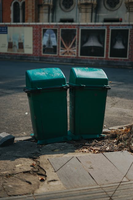 Two green plastic rubbish bins with hinged lids are positioned side by side on a sidewalk curb, close to the edge of a paved pedestrian area. The bins have a smooth, slightly glossy surface with visible black handles on the lids for easy opening. They are situated on a small patch of earth and concrete, with some scattered dried leaves and debris around the base. In the background, there is a street scene featuring a brick building with large windows and decorative architectural details, partially obscured by a fence or wall with posters or paintings. The environment appears to be an urban area, possibly in Richmond, where private waste collection services like Rubbish Collection Richmond provide alternative rubbish removal options. The scene is illuminated by daylight, casting soft shadows and highlighting the clean, functional appearance of the bins, which are ready for waste disposal or collection as part of a local or independent rubbish removal route.