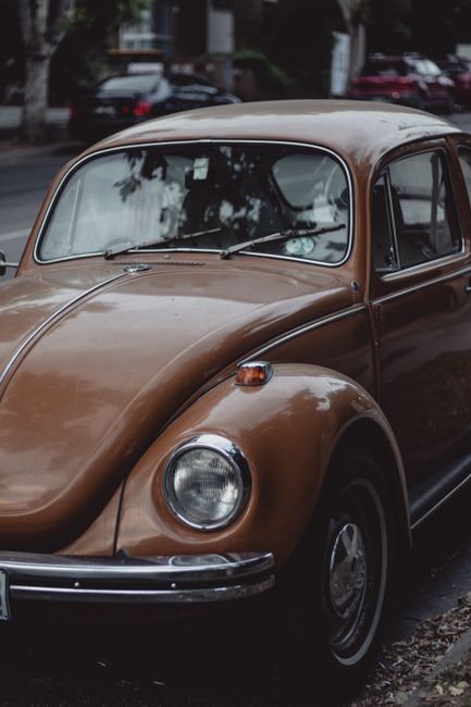 A vintage brown Volkswagen Beetle parked on a street with a background of blurred trees and residential buildings. The car's rounded body and distinctive front features, including circular headlights and small turn signals on top of the front fenders, are clearly visible. The vehicle's surface appears slightly worn, with some age-related patina and minor surface imperfections on the metallic finish. The windshield reflects nearby trees and sky, with the wipers resting at the base. The car is positioned close to the curb, with part of the pavement and a small pile of dry leaves visible underneath. Surrounding the vehicle, other cars can be seen parked along the street, indicating a typical neighborhood setting. This scene subtly reflects the context of classic vehicle maintenance and private collection or disposal of older cars, aligning with the themes of alternative waste handling and vehicle recycling services provided by Rubbish Collection Richmond.